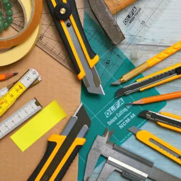 assorted type and size utility cutters on clear and green olfa measuring tool near adhesive tape rolls